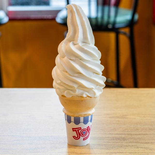 A tall, swirled soft-serve vanilla ice cream cone in a "Joy" branded paper holder on a wooden table.