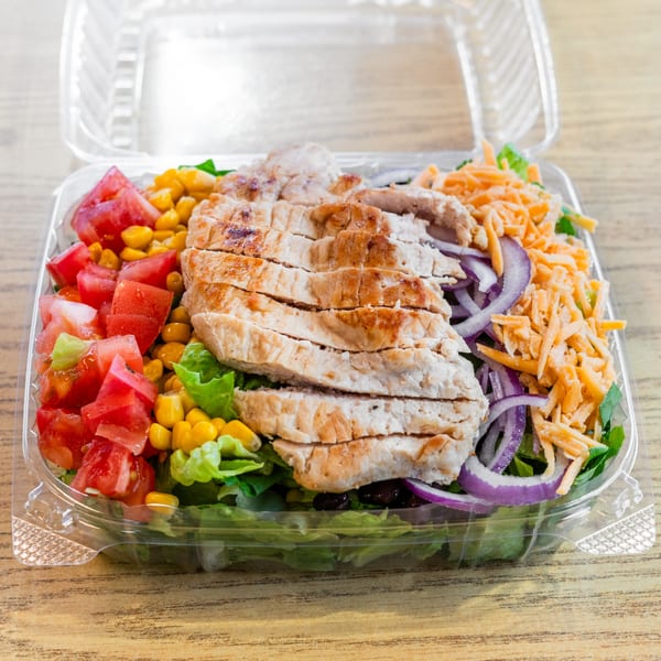 A grilled chicken salad with tomatoes, corn, red onion, and shredded cheese in a plastic to-go container.