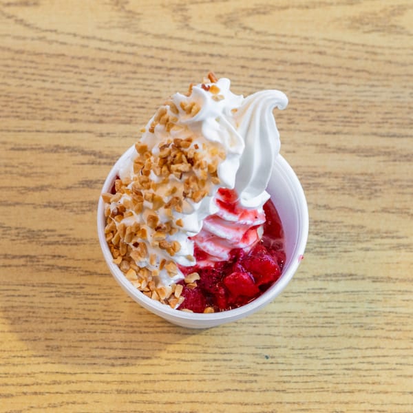 A white cup of strawberry sundae with whipped cream and chopped nuts on a wooden table.