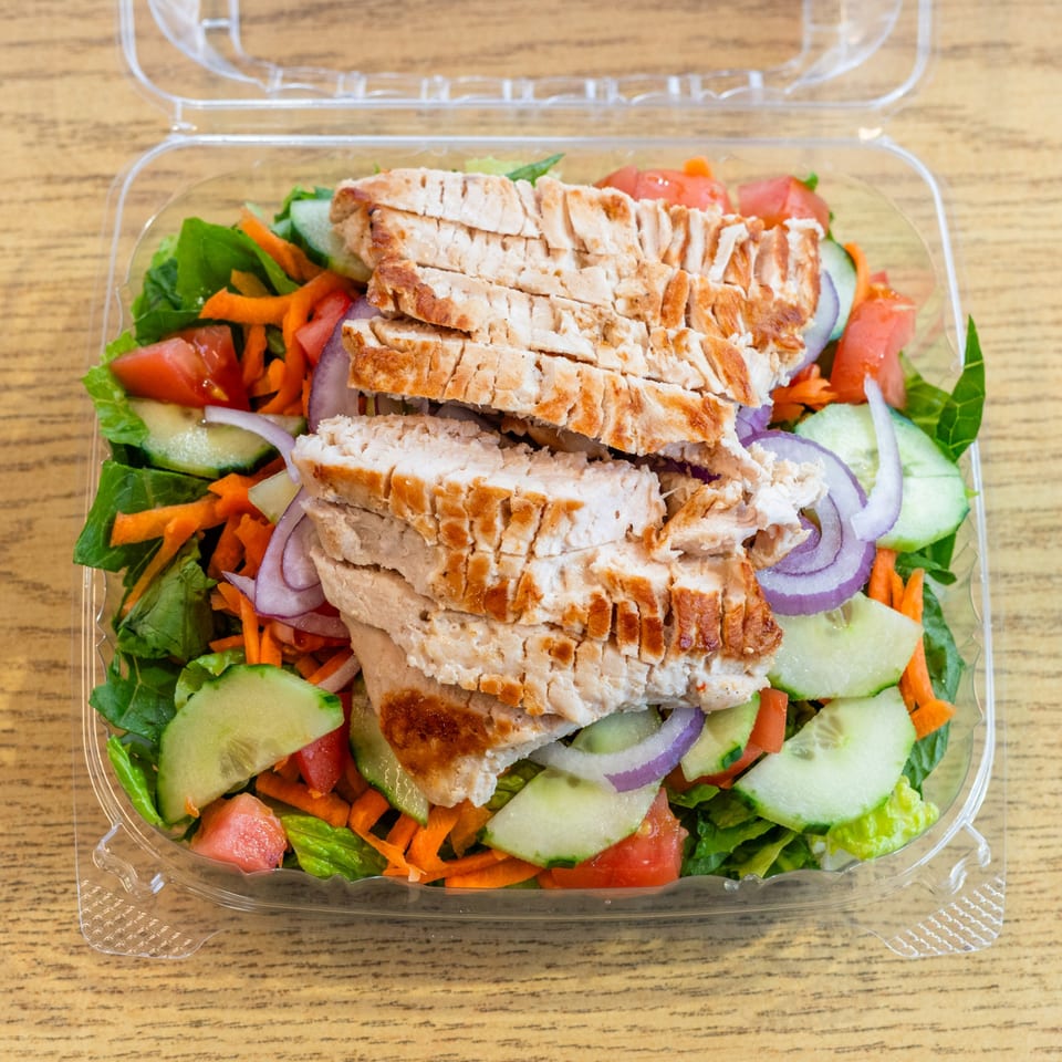 A grilled chicken salad with lettuce, tomatoes, cucumbers, carrots, and red onions in a plastic container.