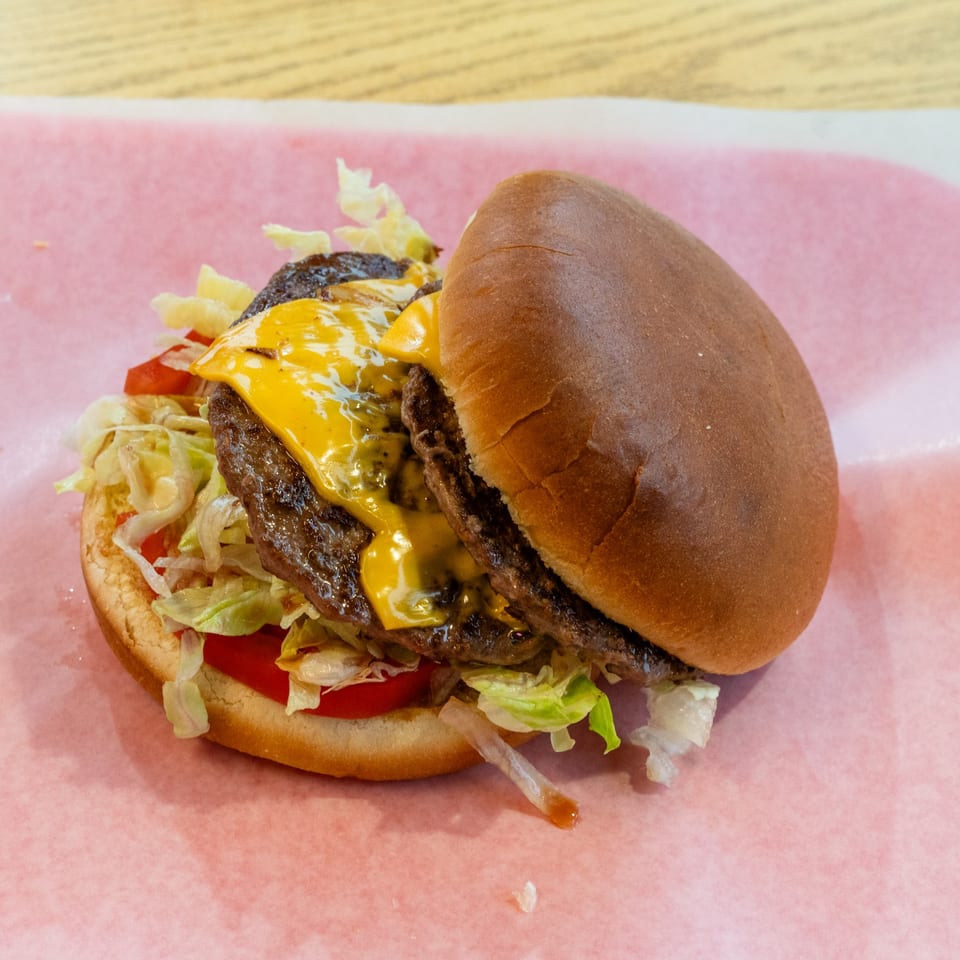 A double cheeseburger with melted cheese, lettuce, and tomatoes on a bun, served on pink paper.